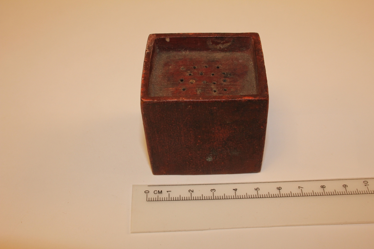 Kubisk boks av tre med 17 små hol i toppen. Botn og topp er senka (topp 8-9mm, botn 4-5mm).