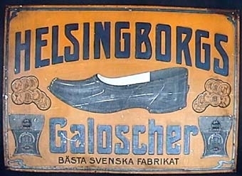 Rektangulär plåtskylt, gul med svart text och en illustration av en galsoch på mitten.
Text: "HELSINGBORGS GALOSCHER
Bästa svenska fabrikat".