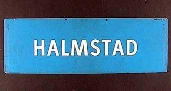 Rektangulär dubbelsidig plastskylt med vit text på blå botten: 
"HALMSTAD".

På andra sidan:
"MALMÖ".