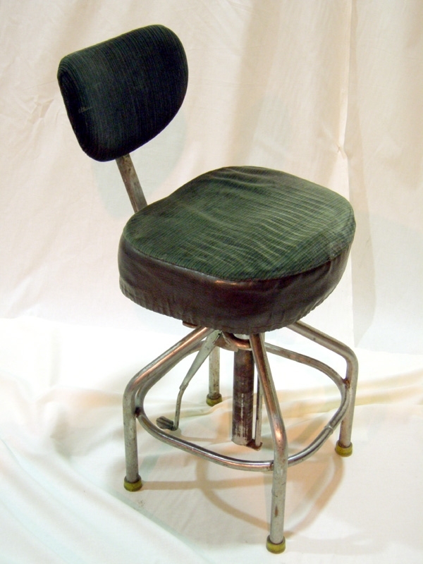 Underrede av stål. Fyra ben av stålrör, på fötterna skor av gul plast. På benens insida en kvadrat av bockat stålrör som fungerar som tvärslåar för stolsbenen, för att göra den stabil. De fyra benen är fastsvetsade i ett ihåligt stålrör igenom vilket ett längre cyliderformat stålrör sitter. På detta sitter i sin tur stolens sits. Det rör som stolen sitter på kan höjas och sänkas med hjälp av en pedal med fjäder. På så sätt kan man enkelt ändra stolens höjd. Stolssitsen består av en tämligen tjockt stoppad sits som är klädd med ett mörkgrönt manchesterliknande tyg. Bred: 435 mm, djup: 365 mm. Sitsen har ett löstagbart överdrag av grönrandigt plyschtyg med galon på sidorna. Sitsskyddet tas på och bort med hjälp av ett resårband som är insytt i skyddets nederkant. Ryggstöd av typisk skrivbordsstolstyp med en för ryggen formad platta, vilken sitter på ett stålrör som i sin tur sitter i sitsens bakkant. Plattan är stoppad och klädd med grönrandigt sammetstyg.
   Har använts som stol till förare av elektrolok. Typen av stol har använts från 1950-talet och används än idag (2007).