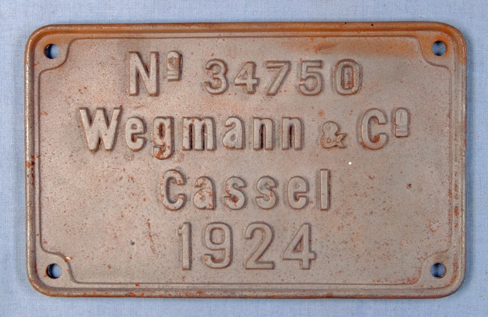 Rektangulär skylt av metall med text i relief. Brunaktig färg.
Text på skylten: "No 34750 Wegmann & Co Cassel 1924".