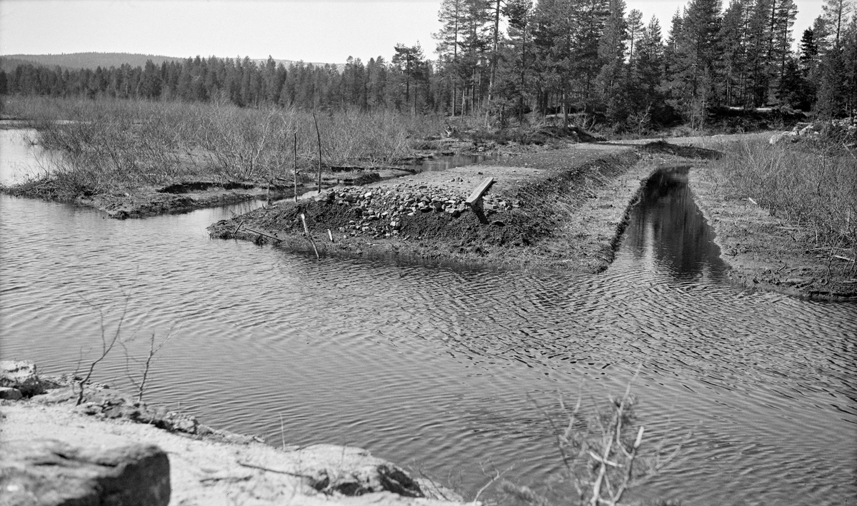 Veianlegg ved Lauervann, mellom Heistadmoen og Skøyen. Fotografiet er tatt på et sted der vegbanen brytes av et stilleflytende vassdrag.  De sto vann også i grøftene på begge sider av veglegemet. Fotografiet skal være tatt i mai 1938, i forbindelse med skogrekruttskolen på Heistadmoen i Sandsvær i Numedal dette året. Soldatene fikk bruke et område omkring Lauervatnet (Løvervann), mellom Heistadmoen og Skrimfjellene, som øvingsområde. Her arbeidet rekruttene med ymse slags hogster, markberedning, såing, skogplanting og grøfting. De bygde nesten 1,7 kilometer med ny veg i tre meters bredde, og oppgraderte bortimot 2,8 kilometer eksisterende veg..
