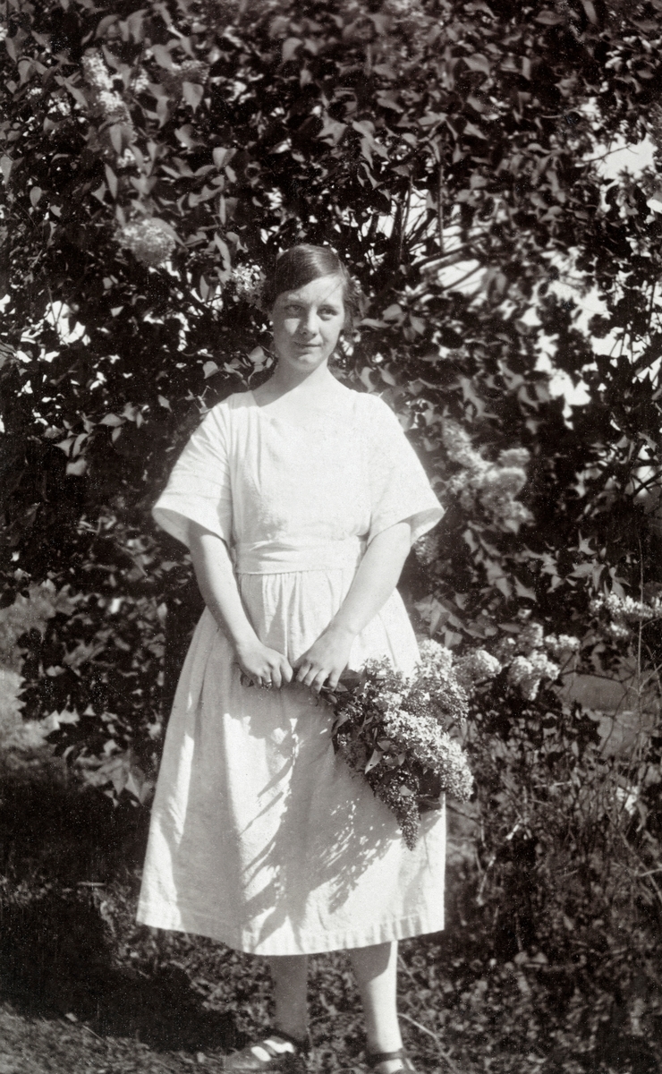 Olga Abrahamsen (1895-1986), kledd i kvit kjole og med en bukett blomstrende syringreiner i hendene.  Hun står antakelig foran busken buketten er hentet fra.  Olga var datter av maskiningeniøren Jänis Eichenberg og hans ektefelle, Līze Kuģiniece Eihenbergs. Faren arbeidet ved et av de tyskbaltiske sagbrukene i Onega.  Olga hadde tatt artium i den latviske byen Mitlava, og deretter fikk hun en kontorpost med ansvar for blant annet utenlandskorrespondanse ved sagbruket der faren arbeidet.  I denne stillingen ble hun kjent med den unge, norske sagbruksbestyreren Egil Abrahamsen.  De to giftet seg i ei urolig tid i Russland, høsten 1917, etter at revolusjonen var i gang og skapte mye uro.  «Fabrikkomitéene» vanskeliggjorde administrasjonen av brukene, og Olgas far ble av den grunn tvunget til å forlate sin stilling i Onega og flytte, etter hvert til hjemlandet Latvia.  Den unge ektemannen Egil fortsatte sin virksomhet som sagbruksleder, i en situasjon som innebar mye usikkerhet, både for bedriften og ham personlig.  Onega var kampsone, med både «kvite», «røde» og vestligeaktører.  Sommeren 1919 måtte Egil, Olga og deres ett år gamle sønn Leif flykte fra rødegardistene som inntok byen, i første omgang til Arkhangelsk, derfra via Murmansk til Vardø.  Derfra reiste Olga og Leif videre sørover, mens Egil reiste østover igjen, blant annet for å betale den gjelden selskapene Russian Forest Industry og Prytz & Co. hadde til russiske banker, i håp om at dette skulle gjøre det mulig å redde noen av investeringene i Kvitsjø-området når situasjonen roet seg.  Olga og barnet bodde i mellomtida først hos Egils søster på Notodden, deretter i et tomt, kaldt sommerhus på Notodden, hvor familiens andre barn Theodor ble født.  I 1920 flyttet familien til Riga, hvor Olgas foreldre bodde.  Lenins pragmatiske økonomiske politikk – «NEP» – gjorde det mulig for Egils norske arbeidsgivere å sende ham tilbake til Onega i 1923.  Der han ble en sentral aktør i et nytt selskap – «Russnorvegules» – der både de vestlige selskapene og den nye sovjetstaten var eierskap.  Olga og de tre sønnene paret nå hadde fått fortsatte å bo i Riga, og dette fotografiet er fra Riga-perioden.  I 1926 flyttet Olga og sønnen etter Egil til Onega.  To år seinere, etter at Stalin hadde kommet til makta, ble de vestlige interessentenes konsesjoner inndratt.  Lederen i det russiske selskapet som overtok virksomhetene hadde imidlertid bruk for Egil Abrahamsens bransjekunnskap og hans språkkompetanse, noe som førte til at det ble ordnet en stilling for ham i en London-bank som arbeidet med østeuropeiske handelstransaksjoner.  Familien bodde der – med unntak av fem krigsår, da de oppholdt seg på hytta i Bamble i Telemark – til Egil ble pensjonist i 1953.  Da flyttet de til Stavern.  I 1970-åra flyttet Egil og Olga til Hamar, der sønnen Theodor var blitt rektor på Katedralskolen.  Begge levde sine siste år i denne byen.