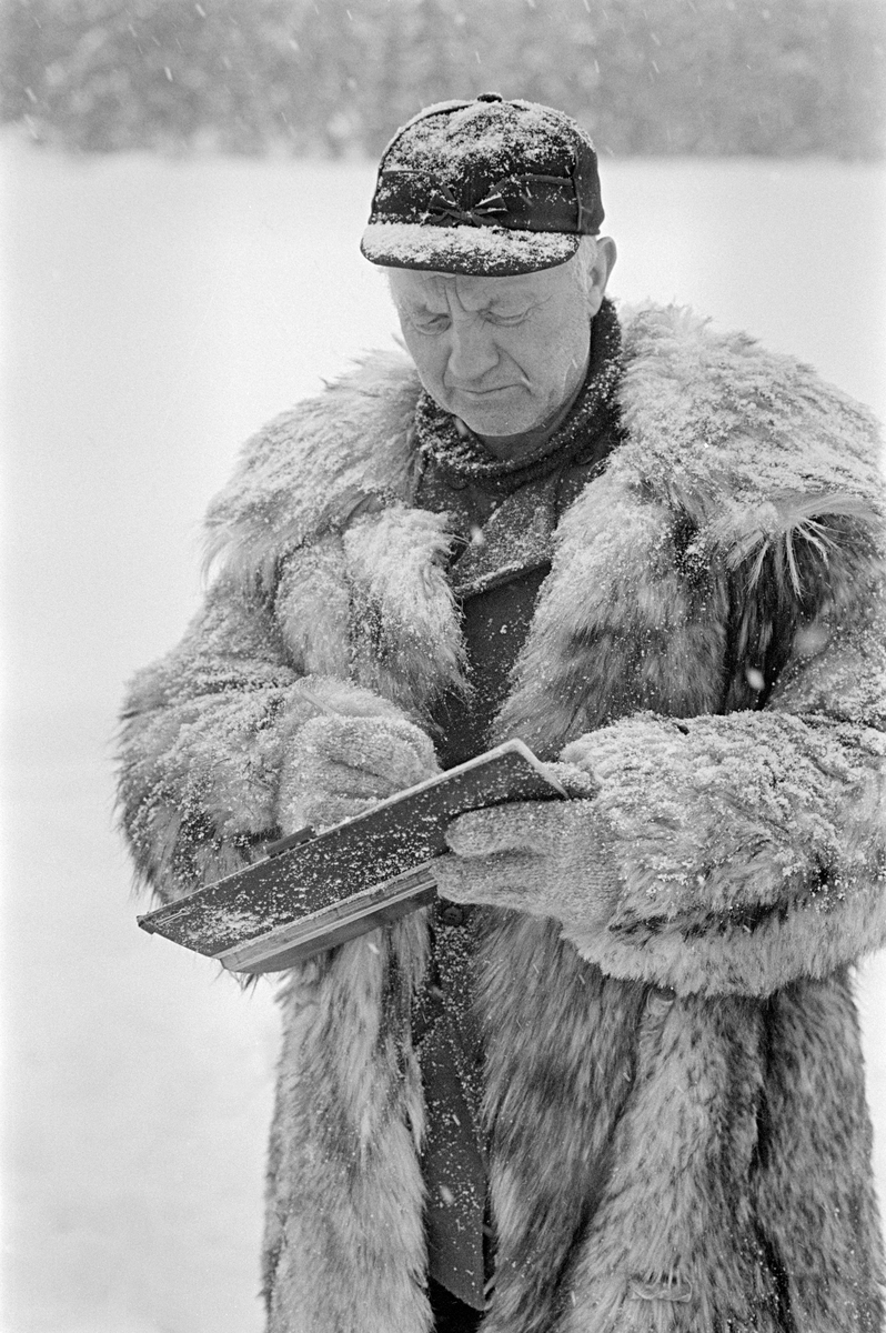Tømmermåling i Nordre Osen (Åmot kommune i Hedmark) i februar 1980.  Fotografiet viser en mann, kledd i ulveskinnspels og med skjoldlue på hodet, med ei stikkbok i hendene framfor seg.  Stikkboka var «notatblokken» der lengde- og diameter på innmålte tømmerstokker ble loggført, med sikte på forestående avregninger mellom tømmerkjøper og skogeier.  Ulveskinnspelsen holdt varmen i måleren under et forholdsvis stillestående arbeid.  Fra gammelt av var det dessuten vanlig av sagbrukenes og treforedlingsbedriftenes målere også var tømmerbetingere, som inngikk kjøpsavtaler.  I sike sammenhenger gjorde de fine kjørepelsene, som ikke var hver manns eiendom, et inntrykk av økonomisk soliditet som skapte tillit i transaksjonene.  Fotografiet er tatt i forbindelse med opptakene til fjernsynsfilmen «Fra tømmerskog og ljorekoie», som ble vist på NRK 1. mai 1981.  Ettersom poenget med denne filmen var å synliggjøre strevet i tømmerskogen i den førmekaniserte driftsfasen, viser den driftsprosedyrer og redskap som bare noen få veteraner fortsatt brukte på opptakstidspunktet.  I 1980 var størstedelen av tømmertransporten egentlig flyttet fra vassdrag til veg, og mesteparten av målinga var samtidig flyttet til kjøpernes tømmertomter.  Dette fotografiet må derfor oppfattes som en rekonstruksjon fra en virkelighet som tidsmessig sett lå minst en generasjon forut for opptaket.