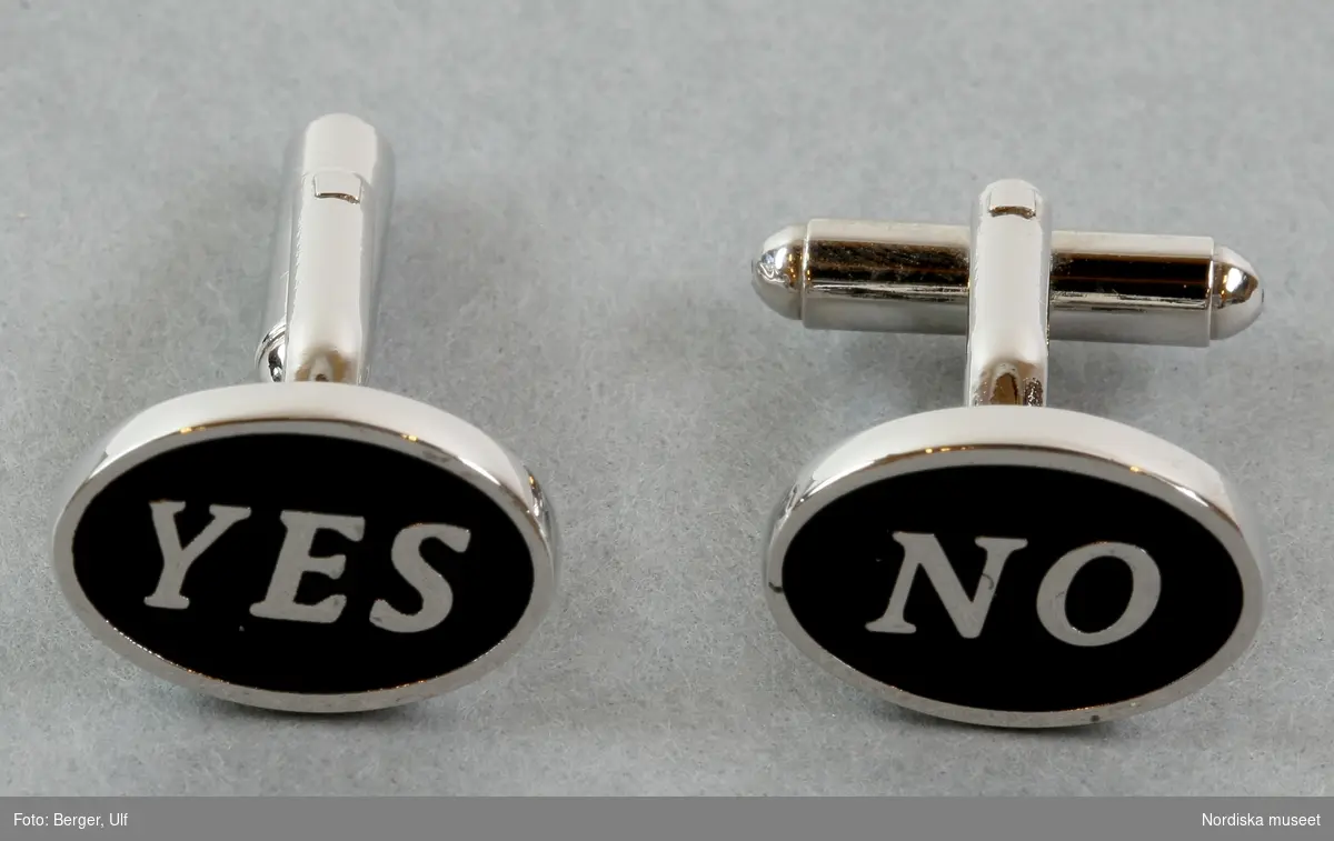 Ett par manschettknappar i metall. Ovansidan är oval (liggande), svart med silverfärgad ram och silvriga bokstäver som säger ”YES” på den vänstra knappen och ”NO” på den högra. Fästanordningen består av två smala metalltenar med en tjockare metallten i mitten som vrids  för att låsa knappen i skjortmanschetten. 
/Elise Wargentin 2017-11