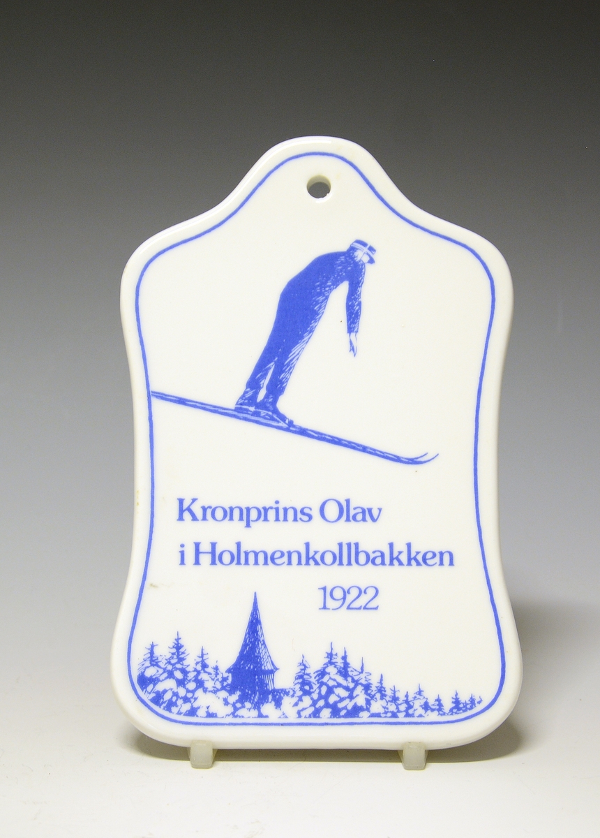 Smørefjel av porselen med hull til oppheng. Motiv av Kronsprins Olav i svev på ski i Holmenkollbakken 1922. Nederst Holmenkollen kappel og granskog.
