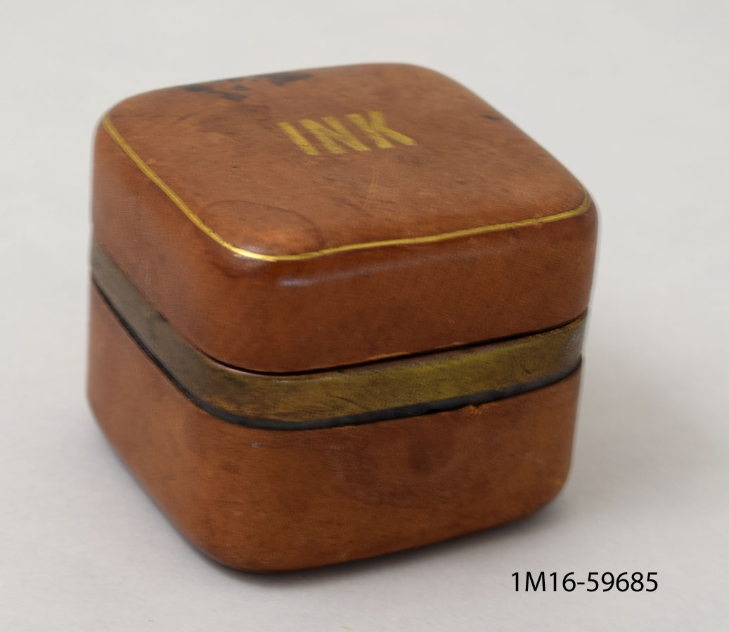 Läderbeklätt bläckhorn av metall beklätt med läder, i formen av en kub. Öppningsbart. Inuti ett bläckhorn av glas. Uanpå är det skrivet "INK".