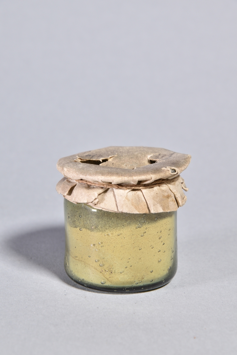 Burk av gröntonat glas, cylindrisk med överbundet lock av papper, fäst med tunt snöre. Innehåller ljust pulver. Locket skadat.