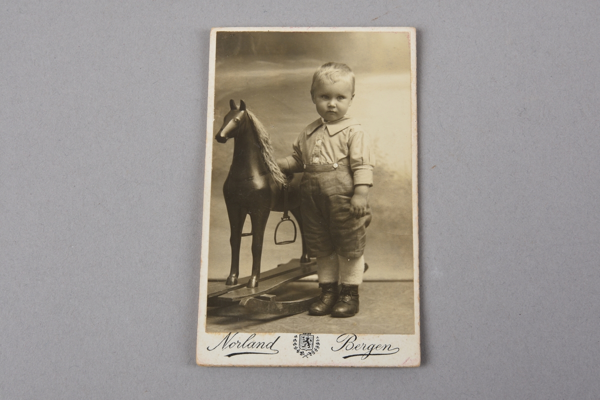 Portrett i svart/kvitt av gut ståande ved siden av ein gyngehest. Guten har på seg skjorte med påknept knebukse, kvite strikkestrømper og svarte snørestøvlar. Hesten har man og stigbøylar.