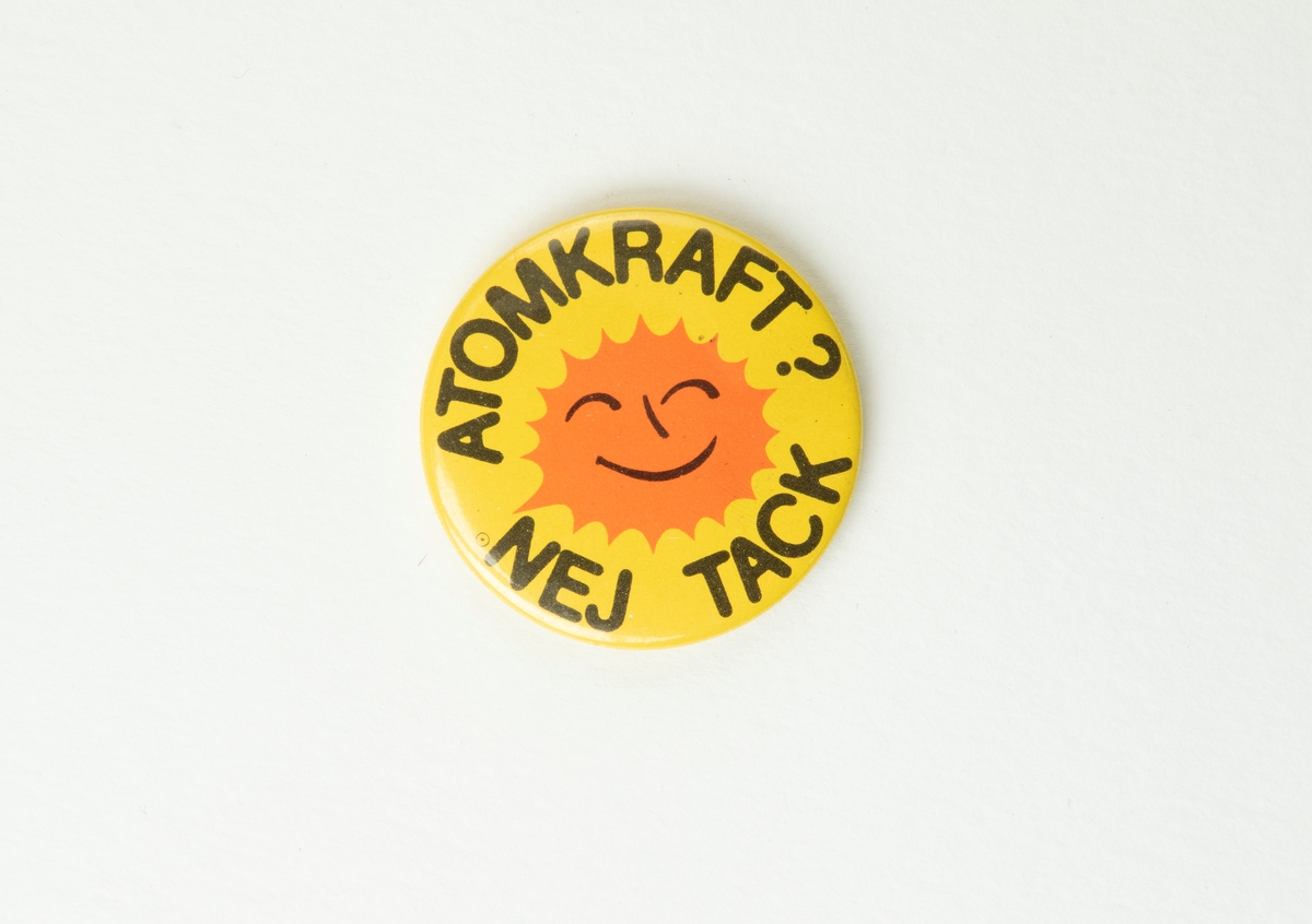Gult rockmärke med en orange glad sol i samt texten "Atomkraft? Nej tack!". På baksidan  en nål som man kan fästa märket med.