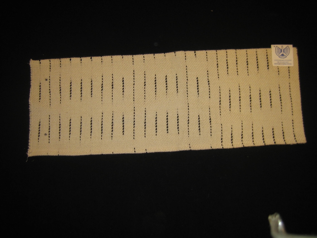 Vävprov i vitt och svart med tvåtrådigt bomullsgarn i varp och entrådig vitt ullgarn i inslag med vitt och svart flamgarn 1,8 cm mellanrum. en lapp fastsatt på tyget med texten: "LÄNSHEMSLÖJDEN SKARABORG SKÖVDE - LIDKÖPING" på ena sidan och texten:"DIAGONAL, BOMULL-YLLE, ANN MARI NILSSON" på andra sidan.