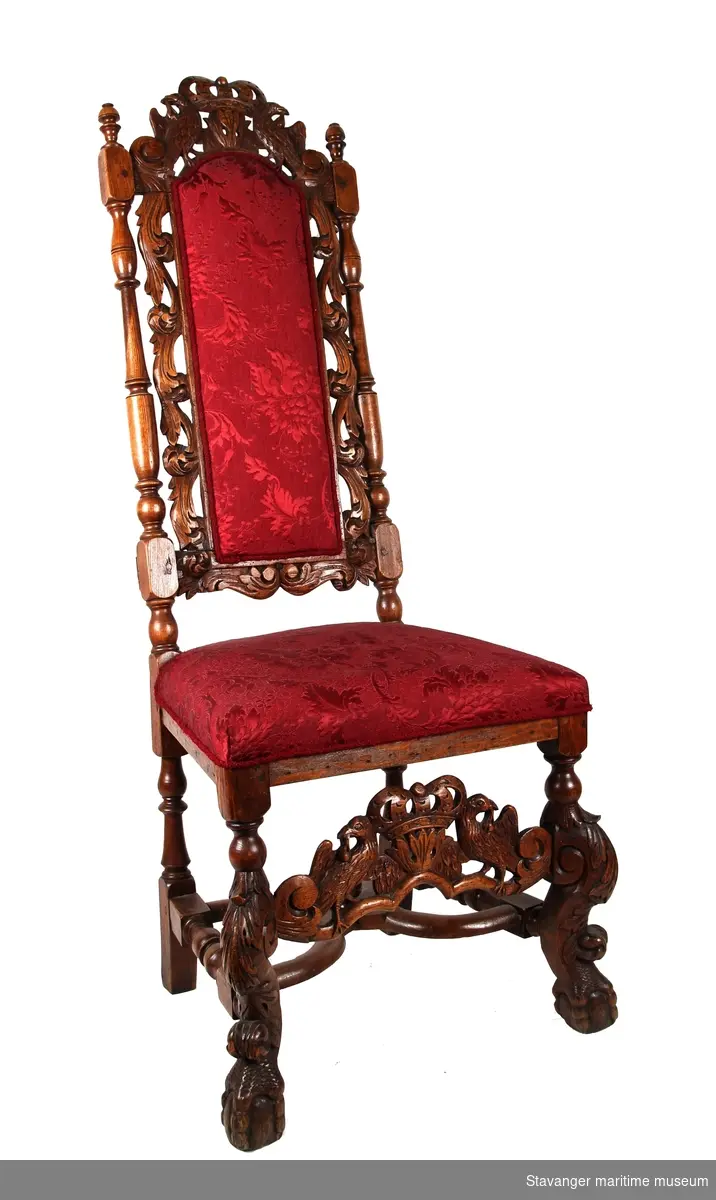 Høyrygget barokkstol av eik med rødt brokadetrekk. Rygg såvel som forben med tilhørende brett er utskåret med dyr og akantusranker.

Sekundært trekk. Ryggen er forsterket  på baksiden i nedrekant.