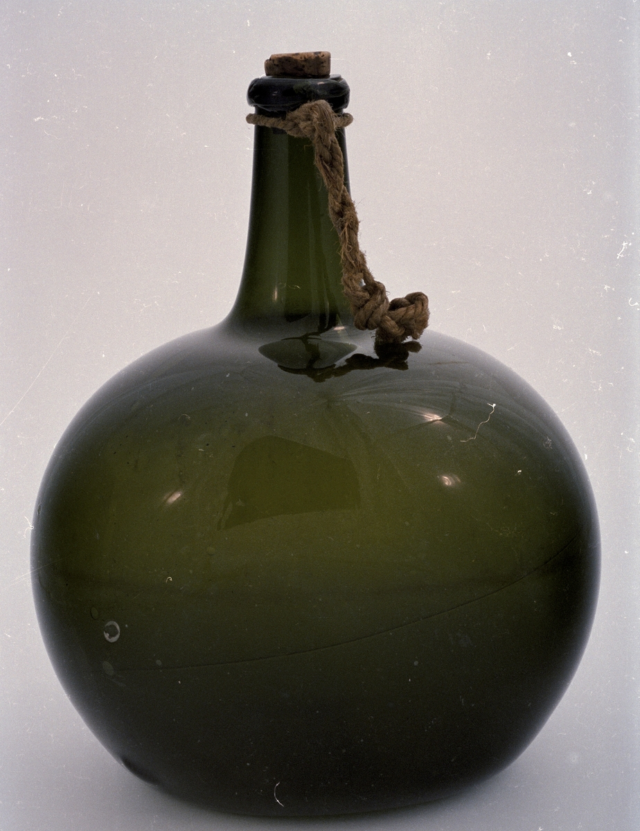 Flaska av grönt glas, klotformad med lätt tillplattade sidor, lång hals med droppskydd samt kork. Vid halsen knutet ett snöre, enligt huvudliggaren för flaskans upphängning.