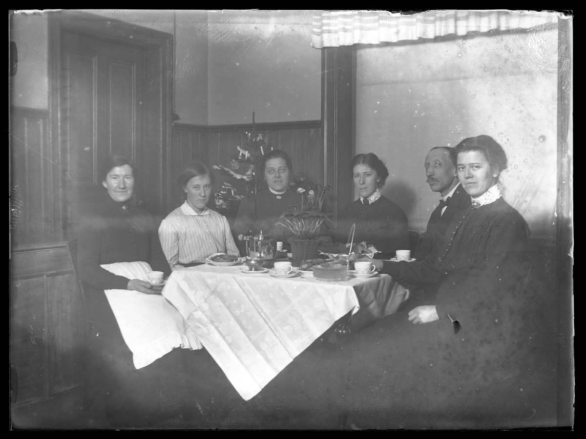 Fem kvinnor och en man sitter vid ett dukat kaffebord och bakom dem står en klädd julgran. I fotografens egna inventeriekatalog står " mor mfl."