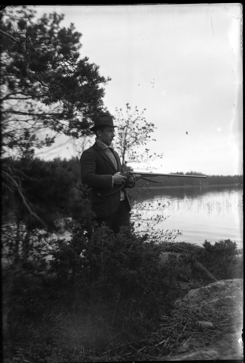 Fotografen själv på andjakt vid Norseskären. Han är klädd i stickad tröja, kavaj och hatt och i händerna håller han en bössa.