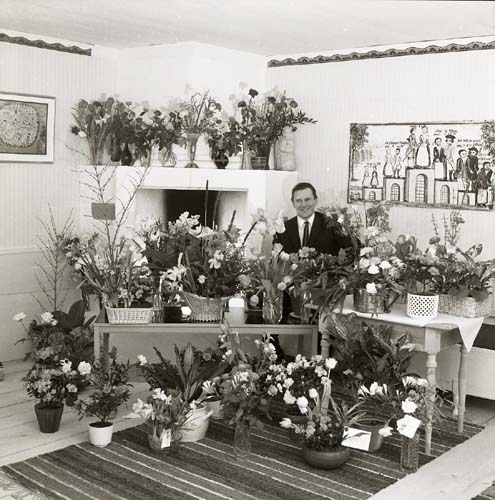 Hilding sitter intill den öppna spisen i ett rum på gården Sunnanåker. Runt honom står massor av blombuketter, 1969. Fotografiet taget på hans 50-årsdag.
