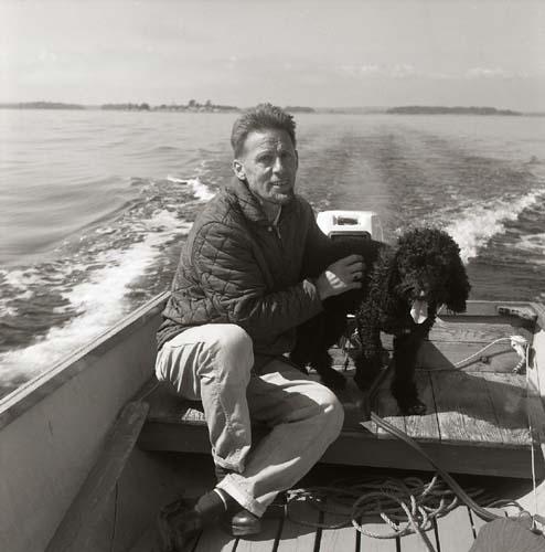 En man sitter vid rodret och kör en motorbåt. Han var mästerlots på Lilljungfruns lotsplats. Intill honom sitter hunden Fabian. Rönnskär, augusti 1969.