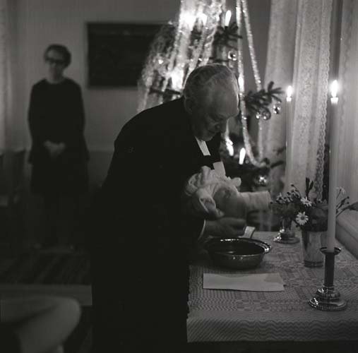 En präst döper ett barn vid ett dopbord. I bakgrunden syns en pyntad julgran, 1970.
