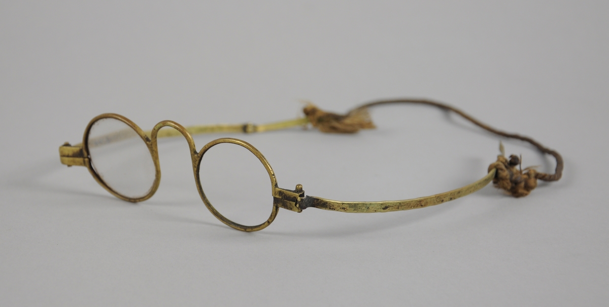 Runde briller med metallinnfatning. Det ene glasset mangler. Stengene kan brettes, og det er knyttet tau til stengerne.