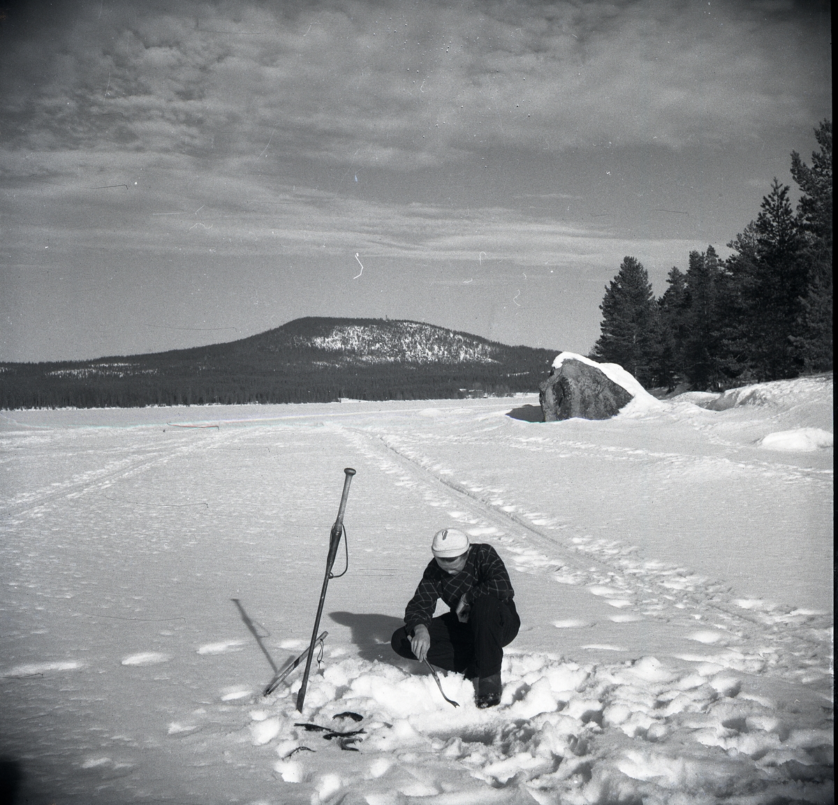 En man som vintermetar på Hölesjön vårvintern 1948. Han sitter på huk vid ett pimpelhål och bredvid sig har han en ispik. Man ser skidpår i snön. Långt i  bakgrunden finns ett berg.