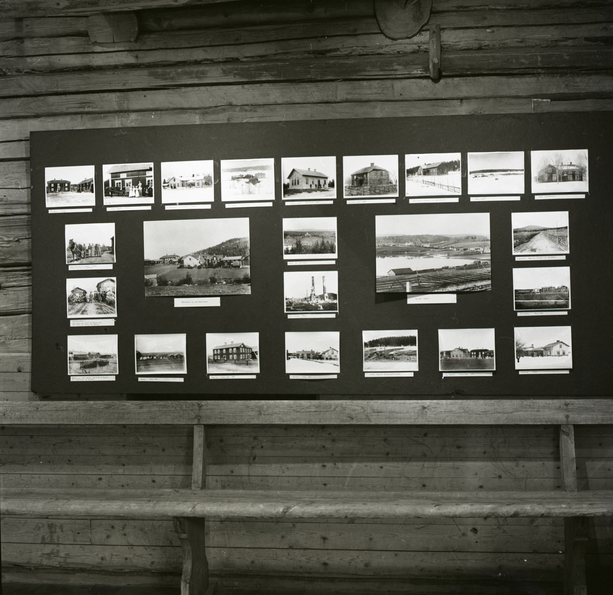 En anslagstavla med fotografier av hus och landsskap ovanför en bänk, 1986.