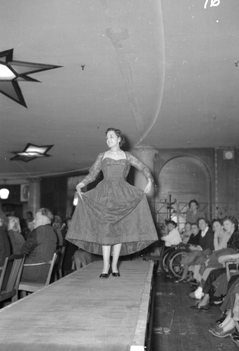 En kvinne på podiet viser fram en kjole med lange ermer. Fra en motevisning på Steen & Strøm fra 7. mai 1954..