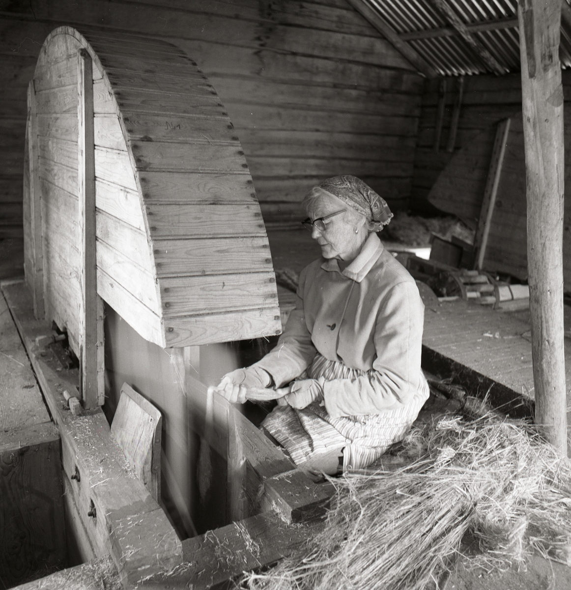 En kvinna sitter vid en linskäkt och bearbetar lin, september 1974. Hon är iförd ett huckle och glasögon.