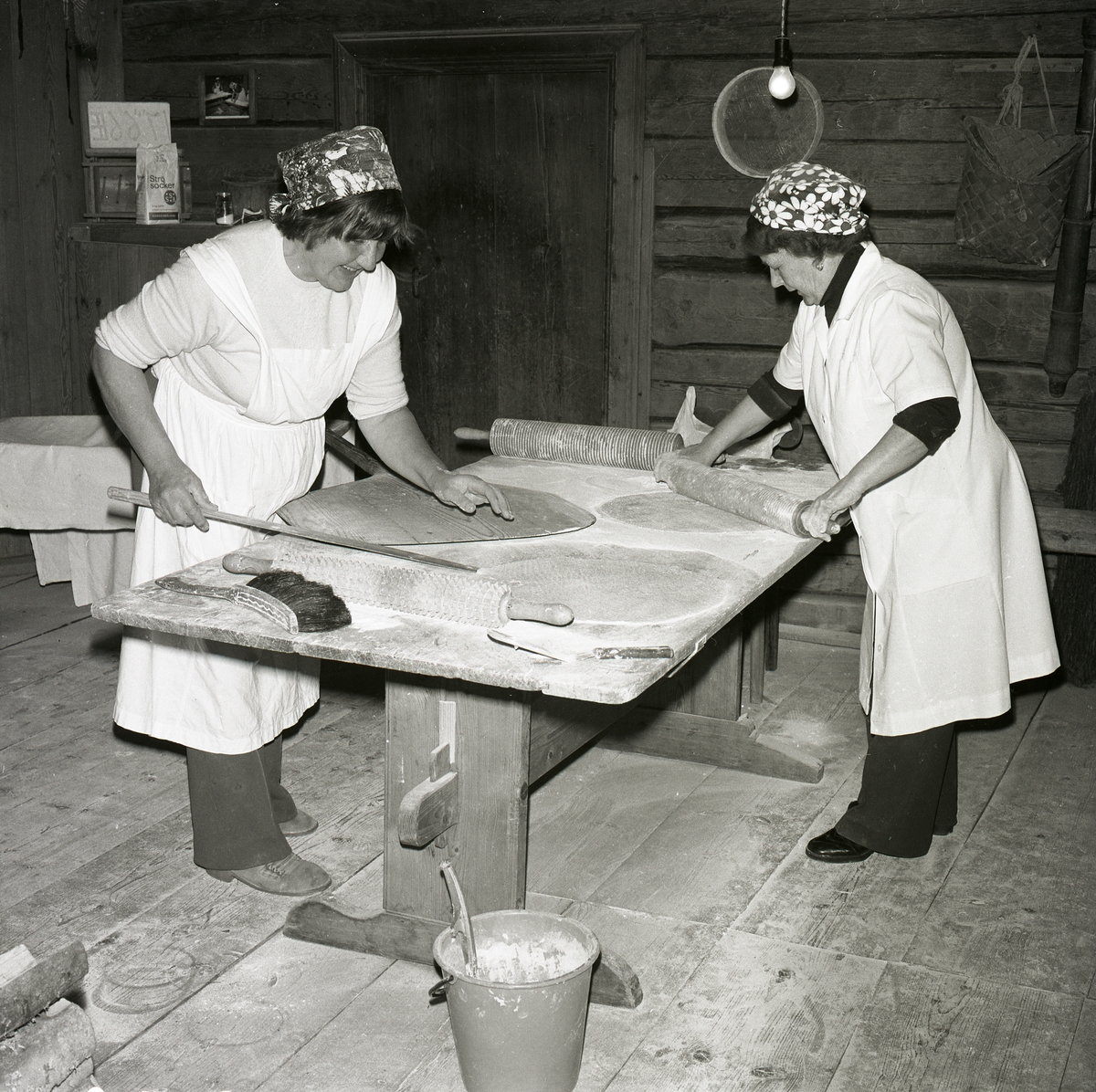 Två kvinnor står vid ett bord och bakar tunnbröd. De använder kavlar, borste, brödpinne och en brödspade som redskap till bakningen. De är klädda i vita täckkläder och huvudbonader, Hembygdsbyn 7 juni 1979.