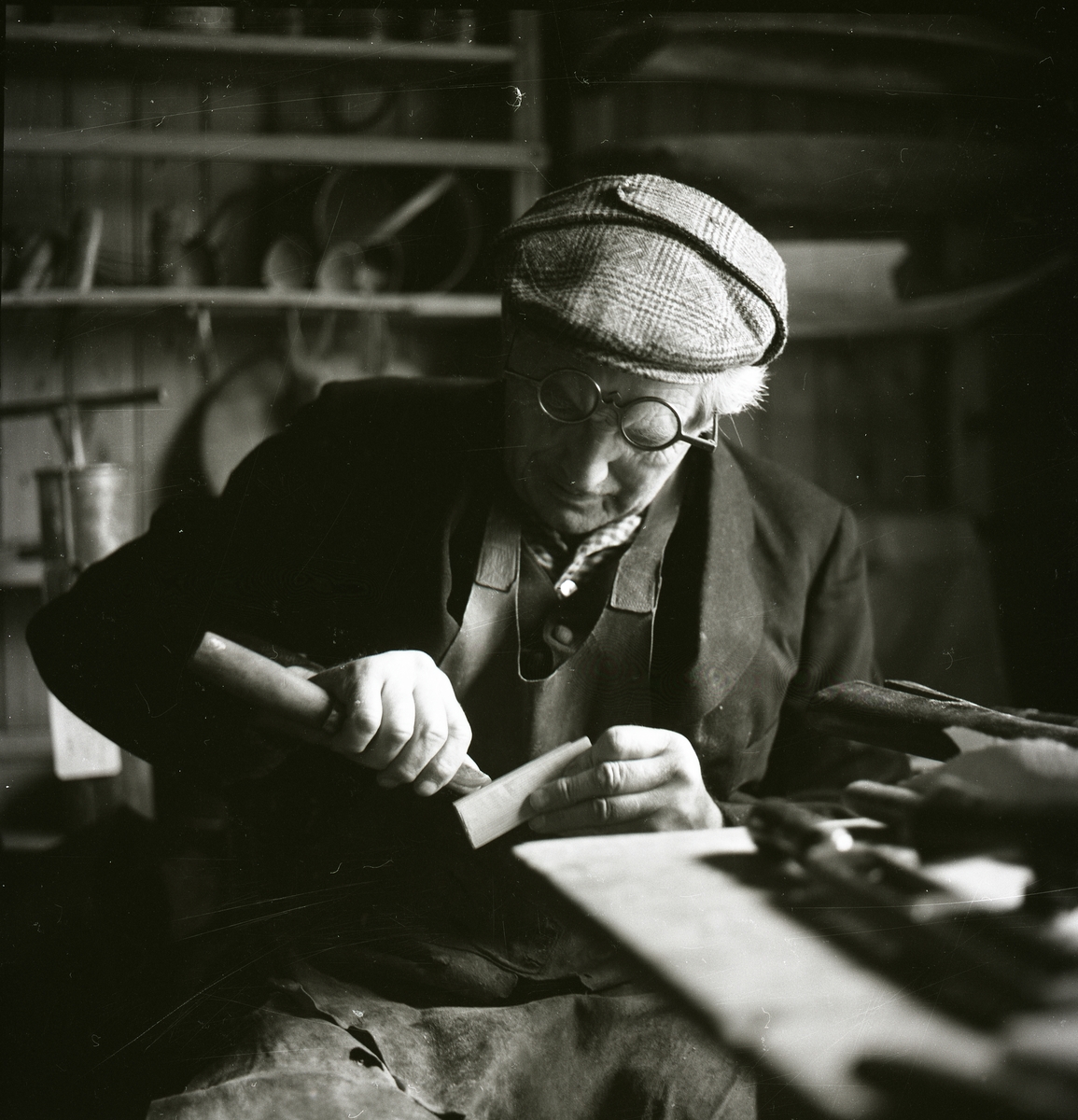 En man iförd ett skinnförkläde, glasögon och huvudbonad täljer ett föremål med en kniv under Gammelvärldens dag vid Hembygdsbyn, 1980 . Bakom honom finns det träföremål på en hylla.