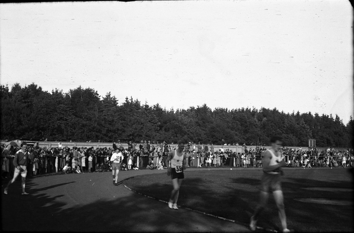 Gångtävling på oidentifierad idrottsarena med tre deltagare i förgrunden och en stor publik längs planket som omgärdar arenan.
(Se även bild EA1199)