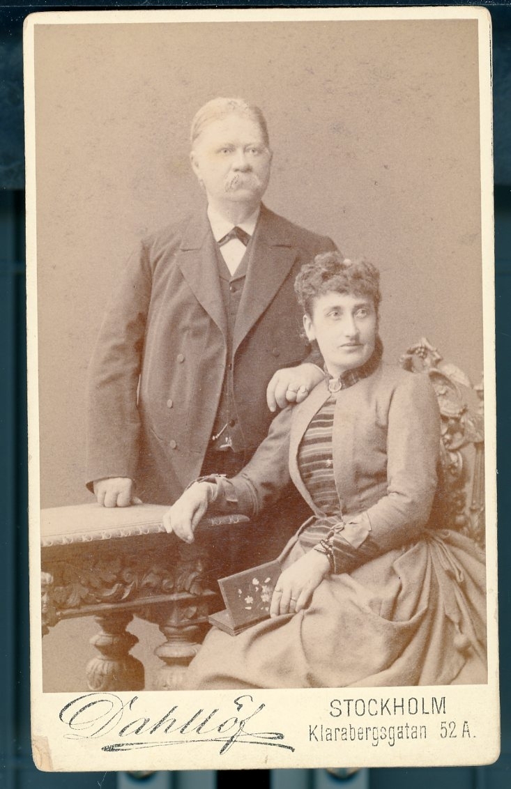 Kabinettsfotografi: J G Hall stående bakom en kvinna (hustrun?) som sitter på en stol. Hans vänsterhand vilar på hennes högra axel.