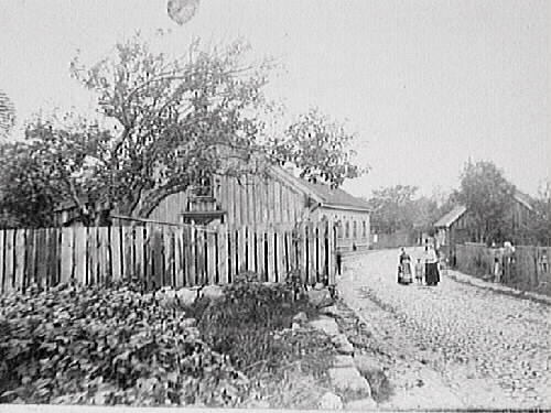 Stadsgata med kullersten utmed ett långt trähus med trädgård på vänster sida och ett staket på höger sida. I mitten står två kvinnor och ett barn på gatan och vid grinden står ytterligare en kvinna.