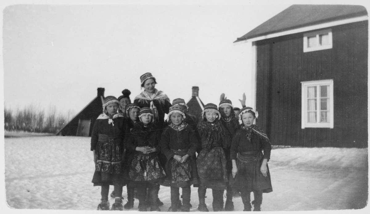 Gruppebilde av samiske barn og en lærer. Bygninger i bakgrunnen.