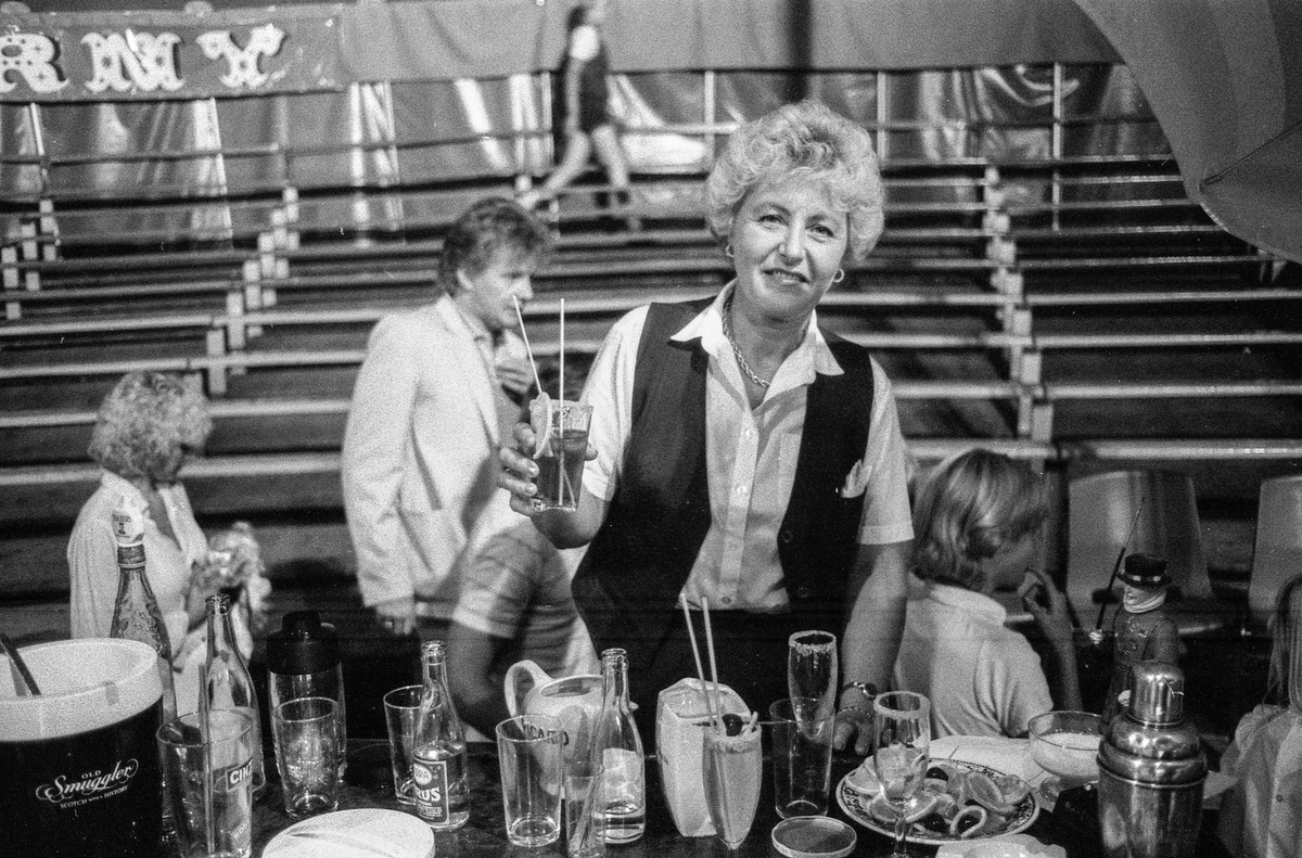 Drøbak-dagene 1982. NRKs hete sirkutelt på Parrstranda i Drøbak.
Mann med paraplydrink. Lill Hansen fra Ski skjenker drinker i baren.