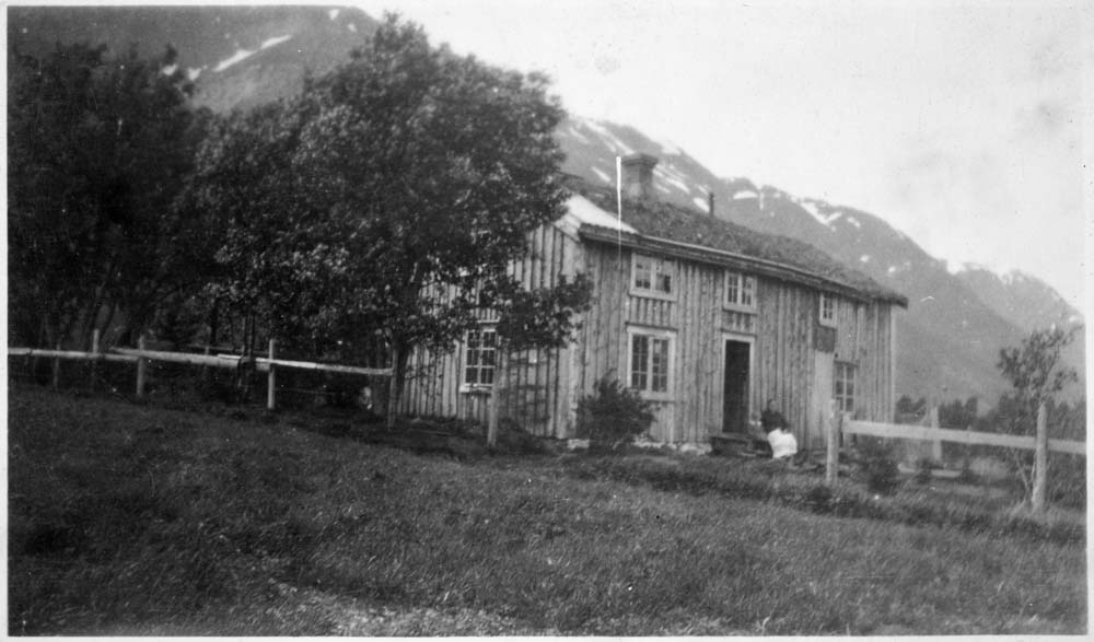 Hus med trær på Øya, Vefsn. Muligens Gudrun Øybakken på trappa ("Gudrun Øybakken" står skrevet bak bildet). Øyfjellet i bakgrunnen.
