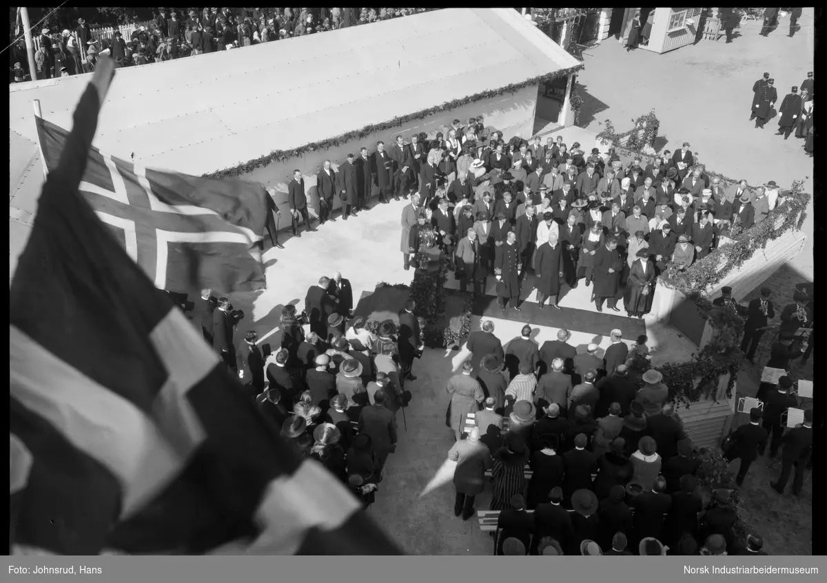 Åpning av Fylkesutstillingen 1922 med besøk av Kong Haakon VII. HM kongen står på rød løper. Korps spiller, jenter i uniform langs veien. Folkemengde rundt hovedarena for åpningsseremonien.
