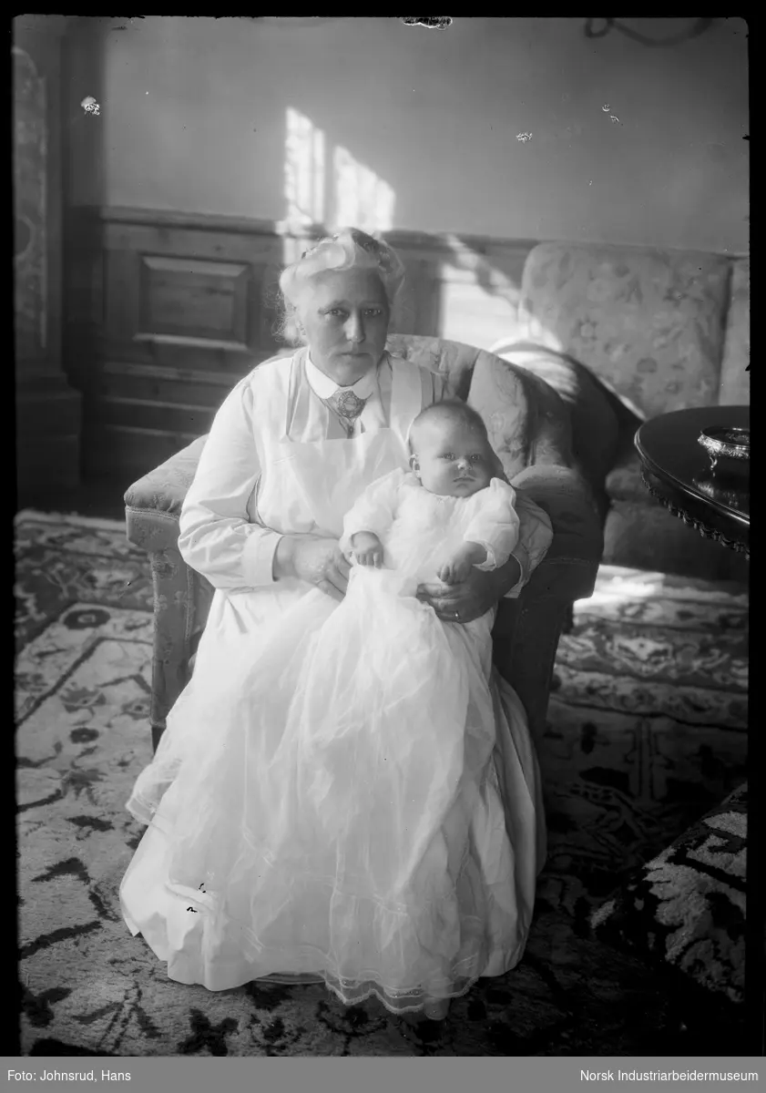 Kvinne sittende med spedbarn i armene. Barnet er kledd i dåpskjole.
