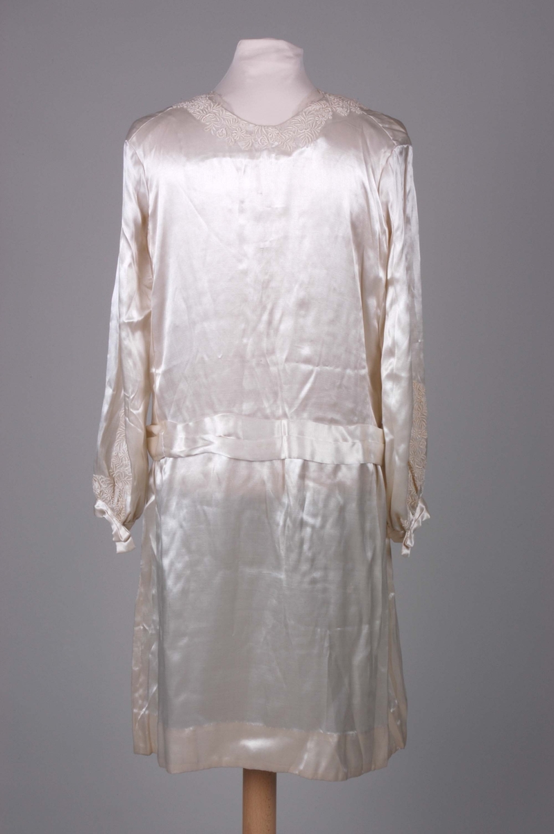 Knelang brudekjole i blank, benhvit silkesateng. Dekorert med innfellinger av tyllblonde.