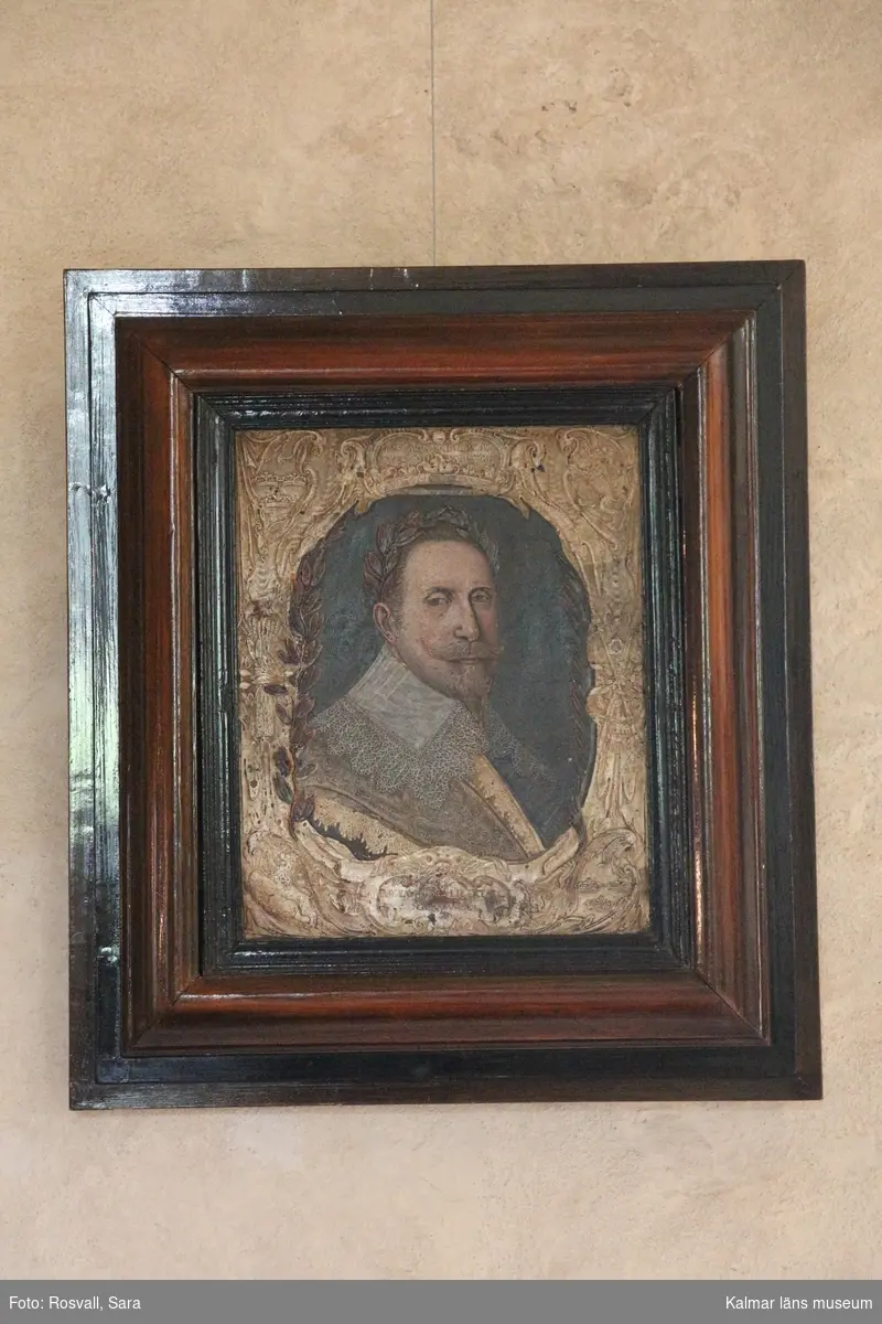Gustav II Adolf porträtterad i bröstbild, face höger. Harnesk och vit spetskrage, kring huvudet lagerkrans. Över harnesket fältbidel, liksom det rika ramverket förgyllt.