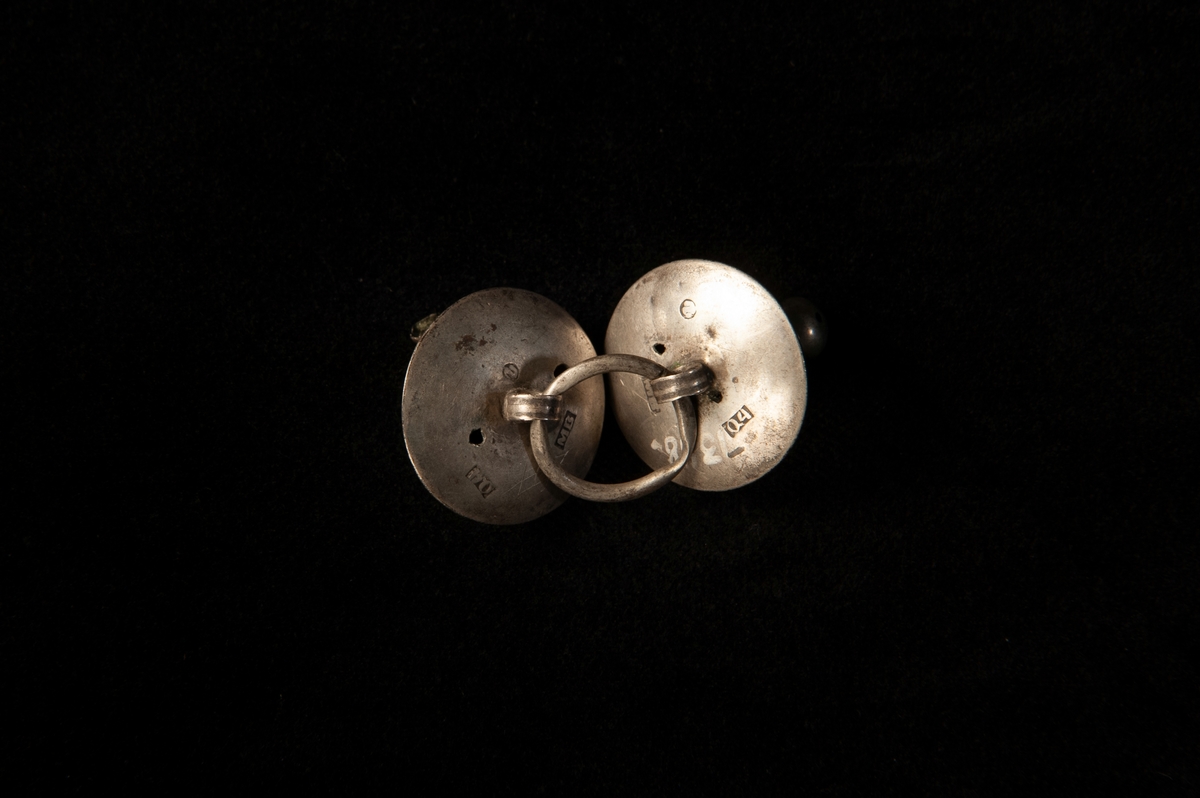 Länkknapp av silver bestående av två kupiga knappar med kula, sammanbundna med ring. Enkel graverad streckdekor.