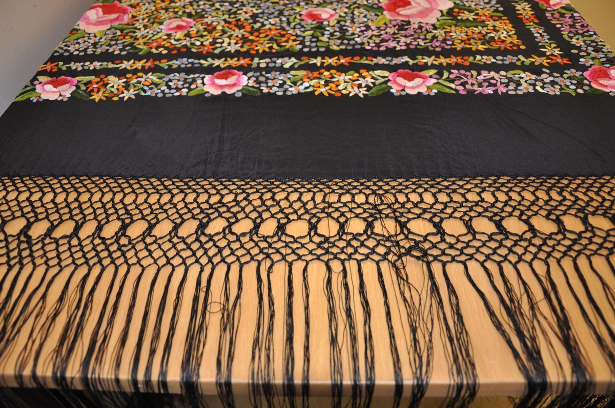 Svart flygelteppe av silke. På midten er det en firkant og to border brodert dekor med motiv av blomster. Rundt teppet er det en kniplet borde med hengene tråder.