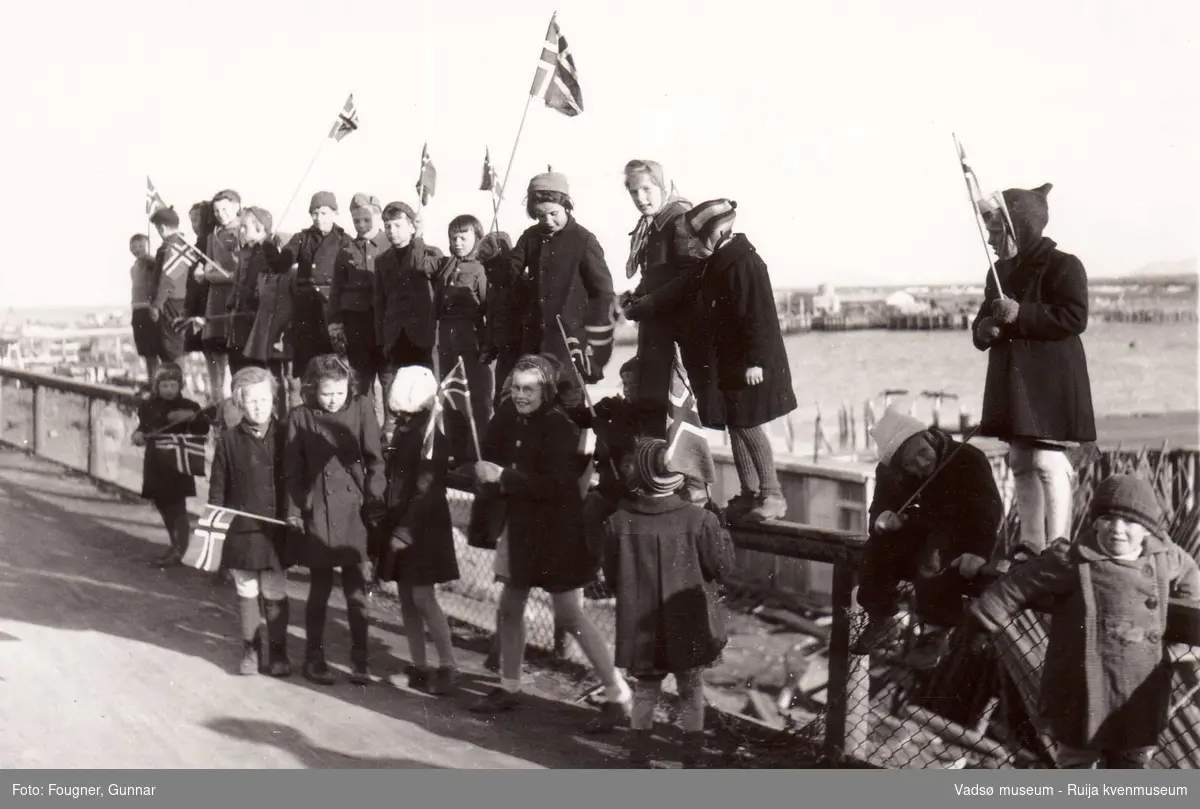 17. mai-tog i Vadsø 1945 med barn som sitter på gjerdet. Vadsø havn i bakgrunnen.