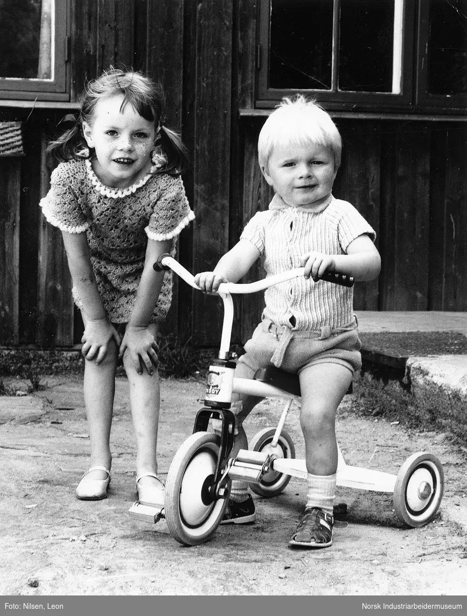 Liten gutt på trehjulsykel, liten jente stående ved siden av.