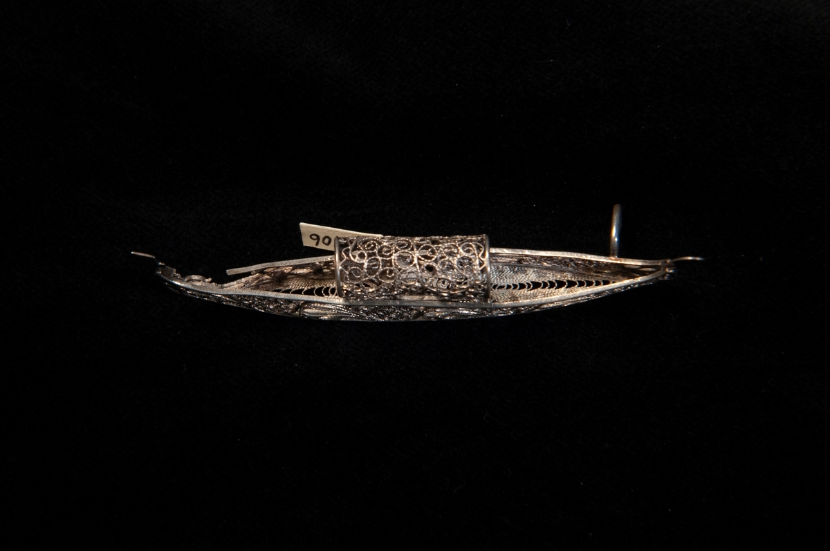 En miniatyrbåt/gondol i filigranarbete i silver. En hängögla finns i skeppets ena del, där den andra öglan troligtvis suttit är filigranarbetet trasigt. Ostämplat.