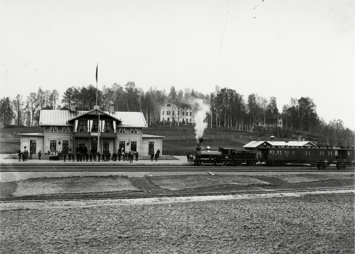 Ångloket MVJ 2 med två vagnar framför stationshuset i Mora. Personal och resande framför, varav tre kvinnor i folkdräkt.