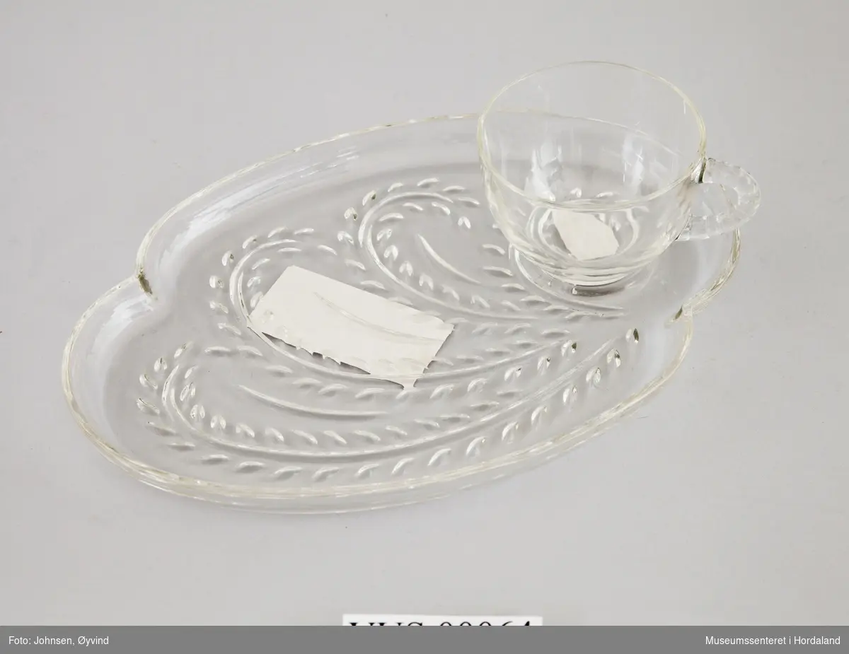 Kopp og tilhørende tallerken i formpresset glass. Tallerkenen er avlang med stråmønster, og har fordypning til koppen. Koppen er glatt med taumønstret håndtak.