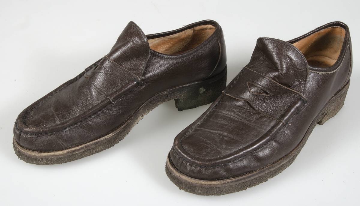Ett par skor (loafers) av ljusbrunt läder med kraftiga rågummisulor. Svarta resårband innanför sleifarna. Överdelarna fastsydda. Maskinsydda. Samhörande med nr. UM 19115-19188.