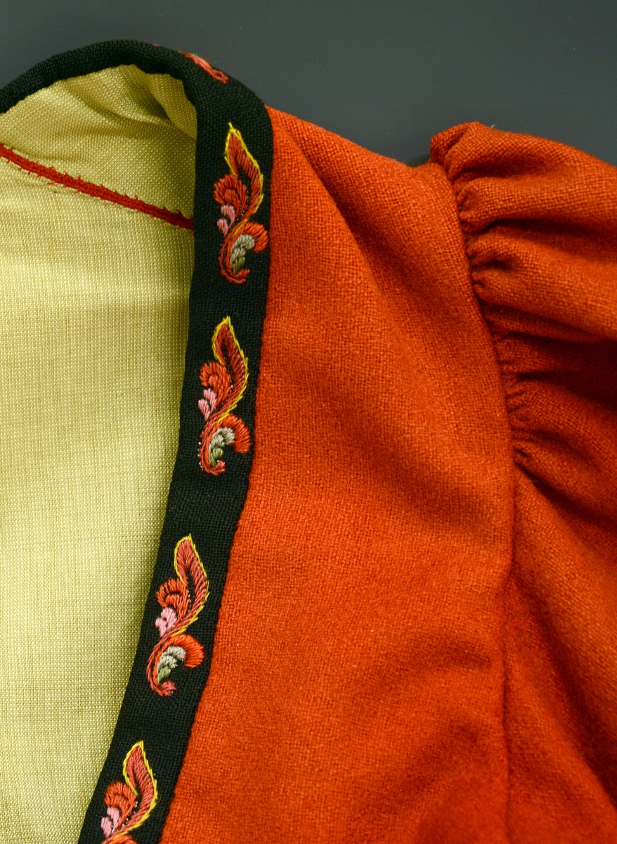 Modellbunad fra Vest-Telemark. TGM-BM-1999:0143.A-D
D. Rød trøye i klede. Foran og på ermene er den kantet med sorte broderte bånd. Broderiene er utført i plattsøm i rosemotiv, fargene er i rødt, rosa, gult, og blått. Trøyen er fòret.