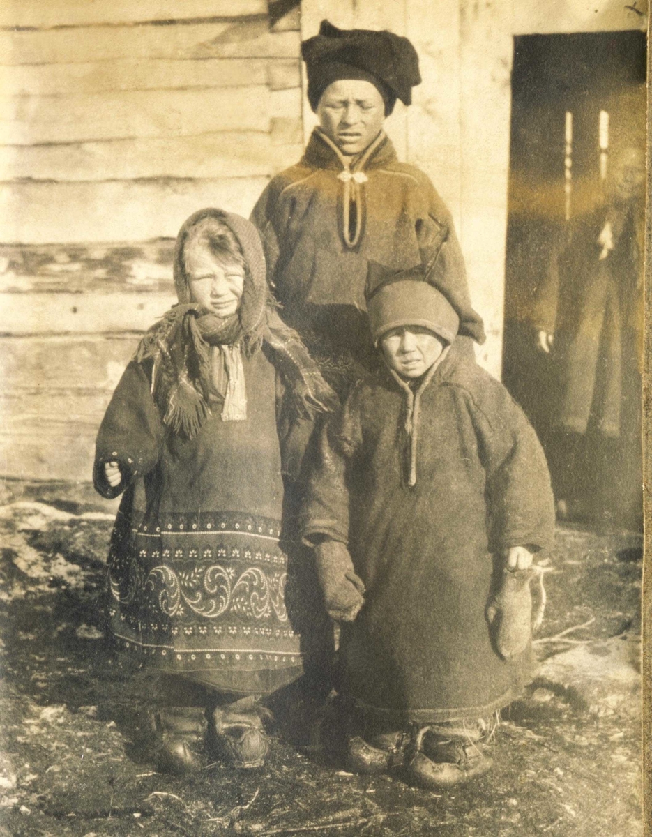 Tre samiske søsken i kofter i Jarfjord. Mor i bakdøra. 1918