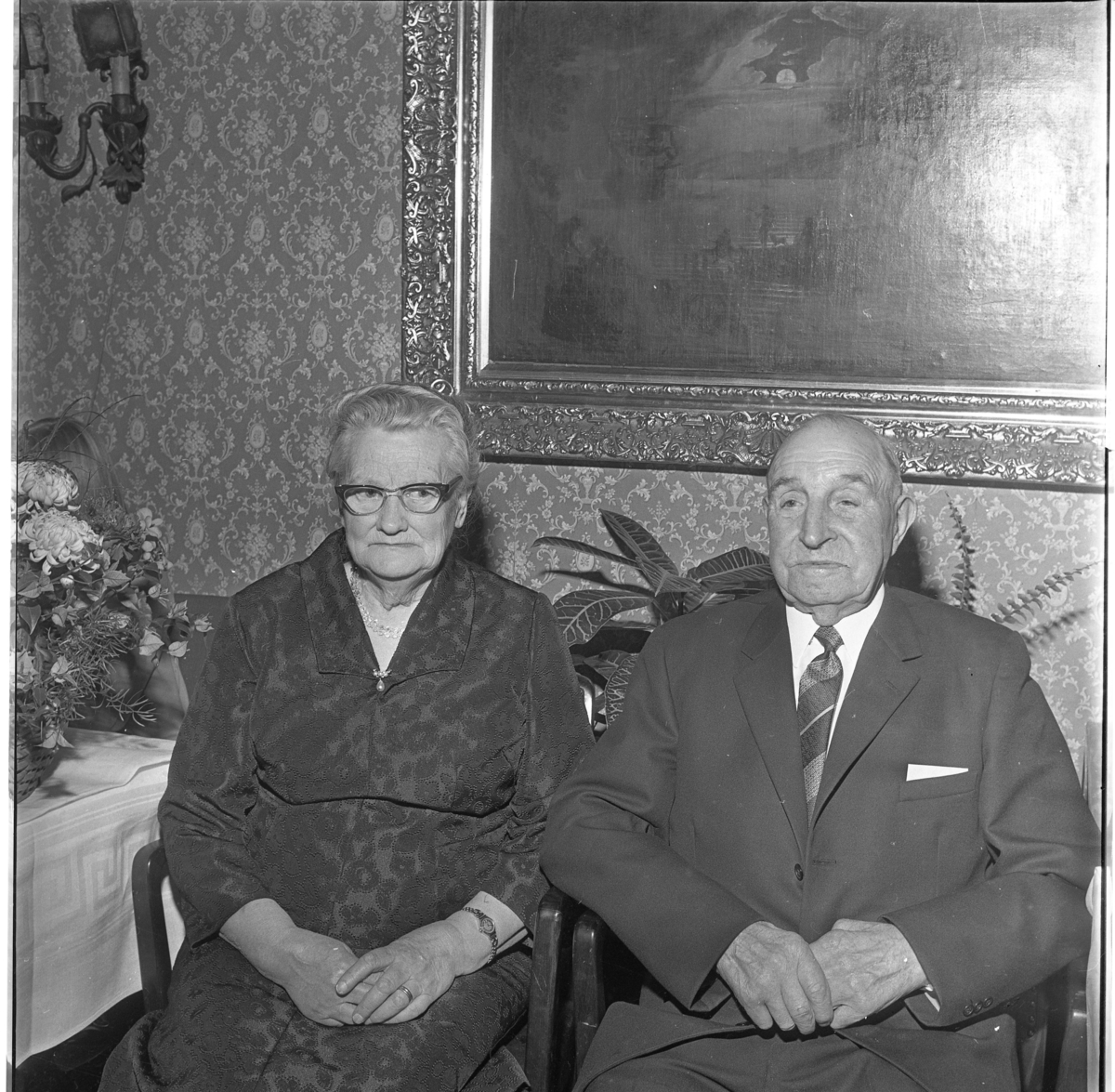 Alma och Josef Larsson i Uppgränna Bultagård firar guldbröllop, 1967. De sitter i varsin stol framför mönstrad tapet med stor oljemålning. Till vänster ett bord där en blombukett skymtar.